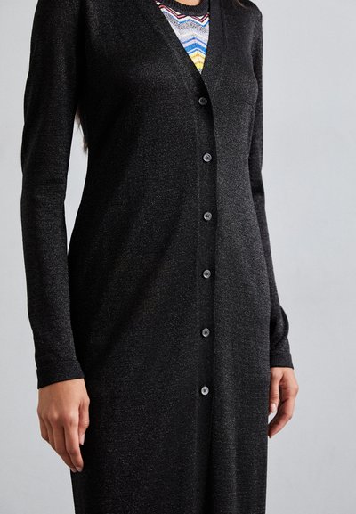 Cárdigan negro con un acabado texturizado, mangas largas, cuello en V y seis botones en la parte delantera. Lleva una capa sobre una blusa de patrón multicolor.