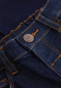 Tessuto di denim blu scuro con cuciture arancioni, che mostra un bottone di metallo con una stella e testo in rilievo vicino a un passante per cintura.