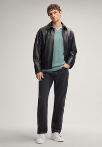 Giacca di pelle nera, maglione in maglia verde, jeans scuri e sneakers bianche. La giacca ha un colletto alto e una texture liscia.
