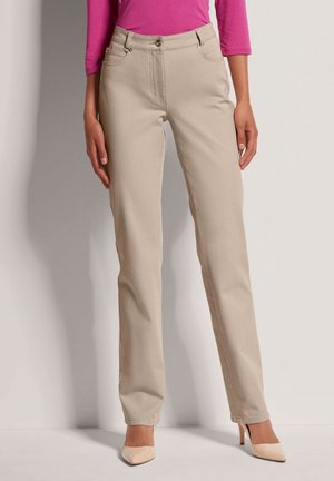 Vrouw draagt beige rechte broek, roze top met lange mouwen en beige hoge hakken, staat tegen een effen achtergrond.
