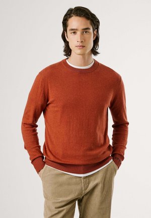 Roestkleurige gebreide trui met een ronde hals, ribbelboorden en -zoom, gedragen over een wit shirt. De textuur lijkt zacht en casual.