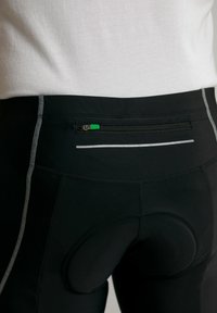 Shorts de cyclisme noirs avec une poche zippée à l'arrière dotée d'une fermeture éclair verte, des détails réfléchissants et un insert rembourré pour le confort. Tissu lisse et extensible.