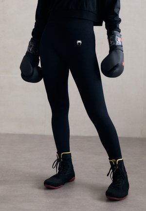 Sorte leggings med en tettsittende design og en hvit logo på midjebåndet, sammen med svarte boksehansker og svarte joggesko med røde såler.