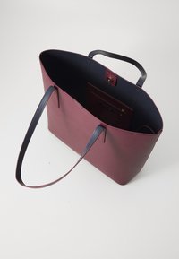 Sac fourre-tout bordeaux au toucher lisse, avec des poignées bleu marine et un intérieur spacieux. Il comporte une petite poche intérieure avec un fermoir à pression.