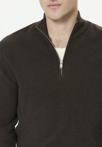 Homme portant un pull à col montant marron foncé avec une fermeture éclair partiellement ouverte, au-dessus d'un t-shirt blanc à col rond, révélant des poils de poitrine et le bas du visage.