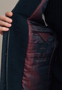 Giacca blu scuro con esterno strutturato. La fodera presenta un motivo geometrico color bordeaux, con un'etichetta etichettata "SIGNATURE" e dettagli per la cura.