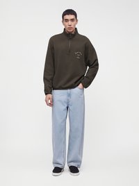 Jeune homme debout portant un sweat-shirt vert foncé à fermeture éclair quart de zip, un jean bleu clair ample et des baskets noires sur fond blanc uni.