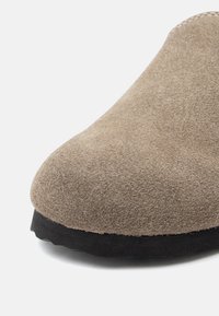 Brun mockaskinn slip-on sko med rund tå, som har en mjuk yta och en svart gummisula för bra grepp. Närbild på övre materialet.