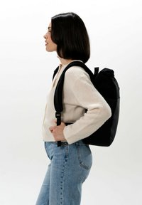 Svart ryggsäck med justerbara remmar, rulltoppstängning och matt yta. Bärs på sidan av en person i en krämfärgad tröja och jeans.