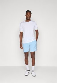 Camiseta blanca de manga corta combinada con pantalones cortos de color azul claro, con un logo. Llevado con zapatillas blancas y calcetines blancos, de pie contra un fondo neutro.