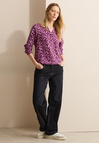 Lila und weiße gemusterte Bluse mit langen Ärmeln, kombiniert mit dunklen Jeans und weißen Sneakers. Die Bluse hat ein V-Ausschnitt-Design.