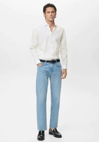 Mand iført hvid skjorte med knapper, lyseblå jeans med sort bælte og sorte loafers, står med den ene hånd i lommen på hvid baggrund.