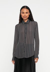 Vrouw met lang krullend haar, gekleed in een doorschijnend zwart overhemd met knopen en verticale witte en roze strepen, gecombineerd met een zwarte geplooide rok.