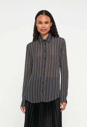 Donna con lunghi capelli ricci che indossa una camicia nera trasparente con bottoni, a strisce verticali bianche e rosa, e una gonna nera a pieghe.