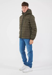 Giacca puffer verde oliva con cappuccio, caratterizzata da pannelli cuciti e chiusura con zip. Indossata sopra jeans blu e sneakers bianche.