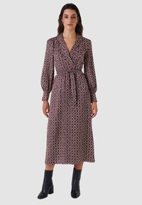 Abito lungo avvolgente color marrone scuro con motivo geometrico. Caratterizzato da scollo con colletto, vita annodata e maniche lunghe voluminose. Indossato con stivaletti neri alla caviglia.