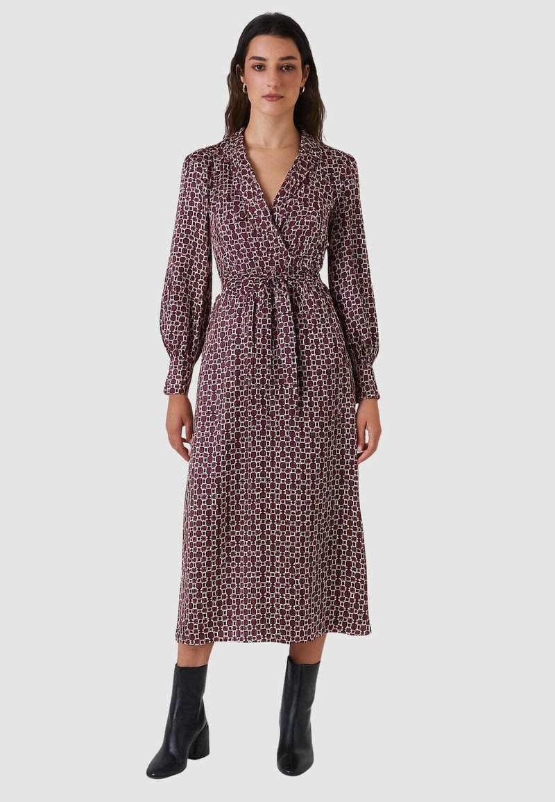 Abito lungo avvolgente color marrone scuro con motivo geometrico. Caratterizzato da scollo con colletto, vita annodata e maniche lunghe voluminose. Indossato con stivaletti neri alla caviglia.