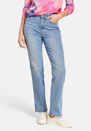 Kvinde iført lyseblå jeans med lige ben, hvide sneakers og en lyserød tie-dye langærmet top med den ene hånd i lommen.