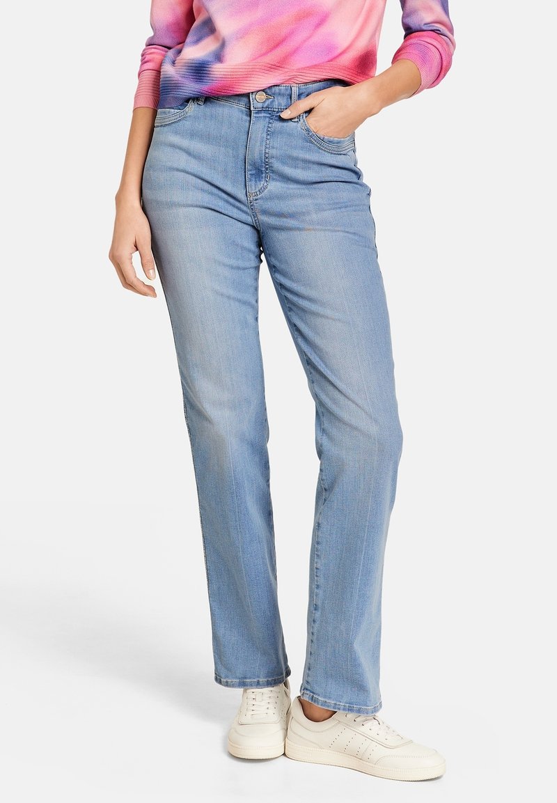 Femme portant un jean droit bleu clair, des baskets blanches et un haut manches longues tie-dye rose avec une main dans la poche.