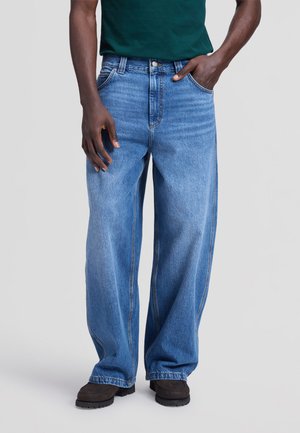 Uomo che indossa jeans blu larghi, scarpe scure e una camicia verde scuro, in piedi con una mano in tasca su uno sfondo semplice.