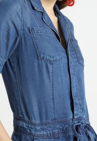 Salopette in denim con maniche corte, due tasche sul petto, vita legata e silhouette aderente in blu medio con dettagli di cucitura visibili.