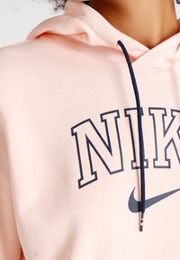 Ljusrosa hoodie med marinblå Nike-text och swoosh-logotyp, som har en marinblå dragsko hängande ner fram.