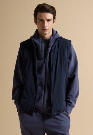 Joven con cabello corto y castaño que lleva una sudadera azul marino con cremallera y un chaleco sin mangas a juego sobre un fondo beige.