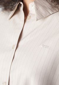 Camicia button-up beige chiaro con sottili strisce verticali, colletto texturizzato e un piccolo logo ricamato sul petto.