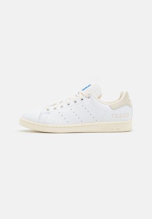 Biały sneaker Adidas Stan Smith z beżową podeszwą i elementem na pięcie, bocznymi perforacjami, złotymi detalami rozmiaru oraz niebieskim logo na języku.