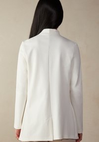 Bílá bunda s dlouhým rukávem a na míru střiženým střihem, vybavená přímým zadním švem a minimalistickým designem, vyrobená z texturované látky.