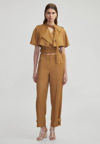 Brown cropped cape jacket with a knot detail, paired with high-waisted brown trousers featuring pockets and cuff accents. Shoes: strappy heels.
