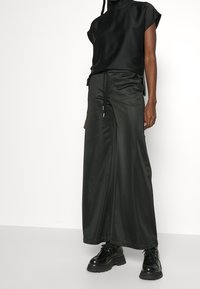 Persona que lleva una camiseta de cuello alto de manga corta negra, pantalones amplios de satén negro con cordón y zapatos de plataforma gruesa negros con cordones, todo contra un fondo blanco.