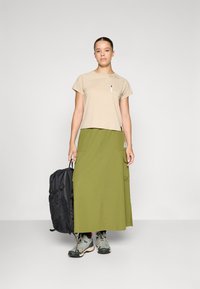 Tricou bej cu mâneci scurte și buzunar în față, asortat cu o fustă lungă de culoare verde măsline, cu buzunare. Rucsac negru și încălțăminte de drumeție gri.