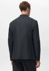 Blazer gris oscuro con corte entallado, que presenta una sola abertura trasera, solapas con muesca y detalles de botones en las mangas.