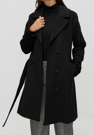 Personne portant un manteau noir croisé sur un col roulé noir et un pantalon à carreaux gris, avec une main dans la poche, sur un fond neutre.