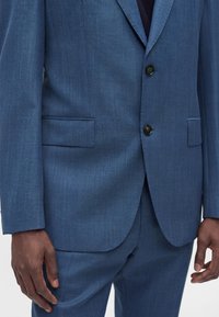 Homme portant un costume bleu texturé avec deux boutons noirs et des poches à rabat, debout, les mains détendues le long du corps.