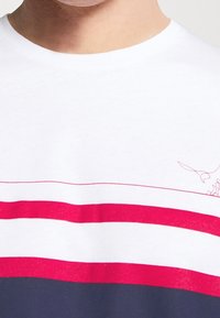 Weißes Baumwoll-T-Shirt mit roten und marineblauen horizontalen Streifen, mit einem kleinen grafischen Akzent nahe dem Ausschnitt. Weicher Stoff, runder Kragen.