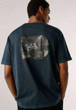 Camiseta de algodón azul oscuro con una imagen cuadrada de un disco de vinilo y un baterista en la parte trasera. Manga corta, corte holgado y cuello acanalado.