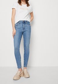 Camisa blanca de manga corta con el texto "vacay", combinada con jeans ajustados de color azul claro y mocasines de plataforma beige. Conjunto sencillo y casual.