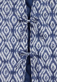 Vuorattu liivi, jossa on sininen ja valkoinen timanttikuvio, suljettavana nauhoilla ja sileällä pinnalla. Koristeltu ommelluilla yksityiskohdilla.