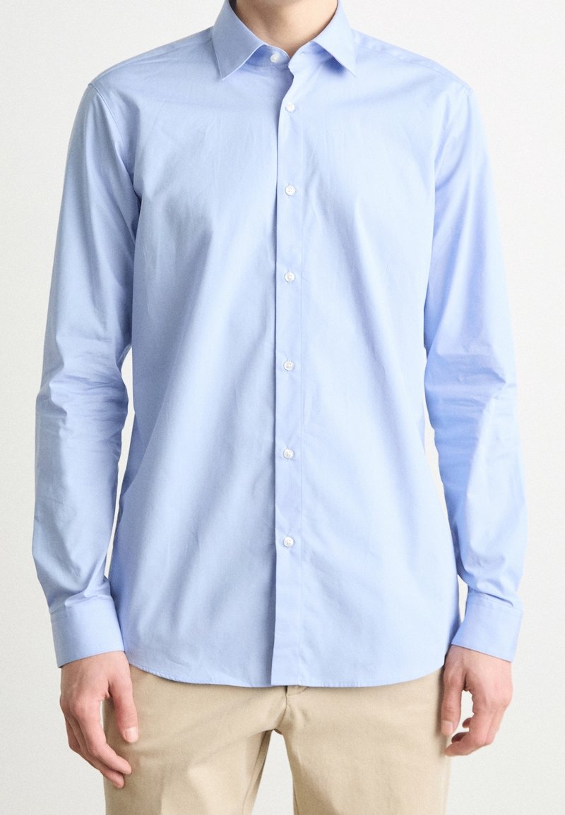 Homme portant une chemise bleu clair à manches longues et un pantalon beige, debout, les bras relâchés sur un fond uni.