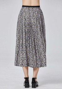 Femme portant une jupe midi plissée avec un motif floral noir, blanc et beige, associée à des chaussures à talons blocs en vernis noir.