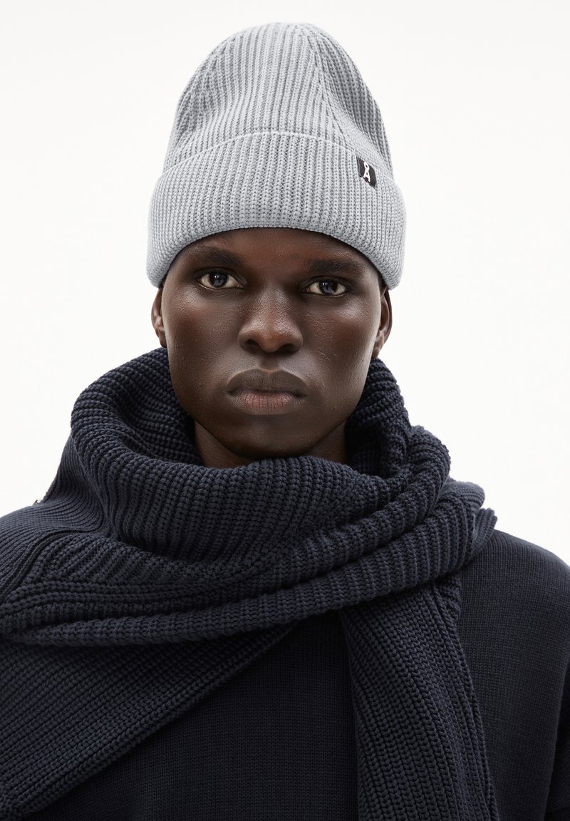 Donkerhuidige persoon met een grijze gebreide beanie en een chunky donkerblauwe gebreide sjaal over een bijpassende trui, recht vooruit kijkend met een neutrale uitdrukking.