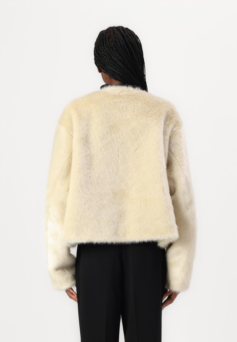 Krótka kurtka z sztucznego futra w kolorze kremowym, charakteryzująca się miękką fakturą, szerokimi rękawami i prostym wykończeniem. Nosi się ją na czarnych spodniach.