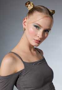 Siva off-shoulder majica s tankim trakama, prilagođenog dizajna. Model ima dvije pletene punđe, odvažnu tirkiznu sjenu za oči i zlatne naušnice u obliku prstena. Glatka tekstura kože.