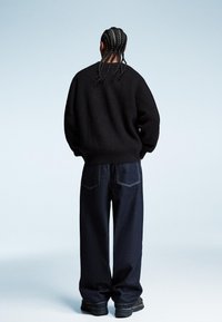 Czarny sweter dzianinowy, luźne niebieskie dżinsy, i czarne buty. Widok z tyłu na tle jasnego gradientu.