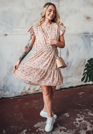 Floral-patterned, beige dress with short ruffled sleeves, gathered waist, and ruffled hem, paired with white sneakers and a small handbag.