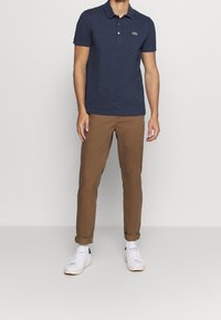 Polo shirt bleu marine avec un col et trois boutons, associé à un pantalon marron fuselé. Des baskets blanches complètent la tenue.