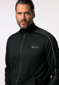 Chaqueta atlética negra con cremallera y cuello alto, con acentos en gris claro y un logotipo en el lado izquierdo del pecho. Fabricada con una tela suave y elástica.