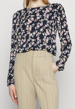 Blusa floreale a maniche lunghe di colore navy con fiori rosa e gialli, abbinata a pantaloni beige a righe sottili. La blusa ha una vestibilità ampia e un colletto rotondo.